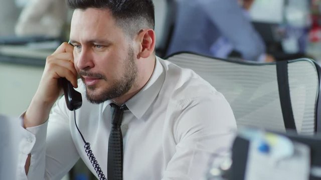 Medium shot of middle-aged man in white shirt and necktie sitting in office and talking on the phone
