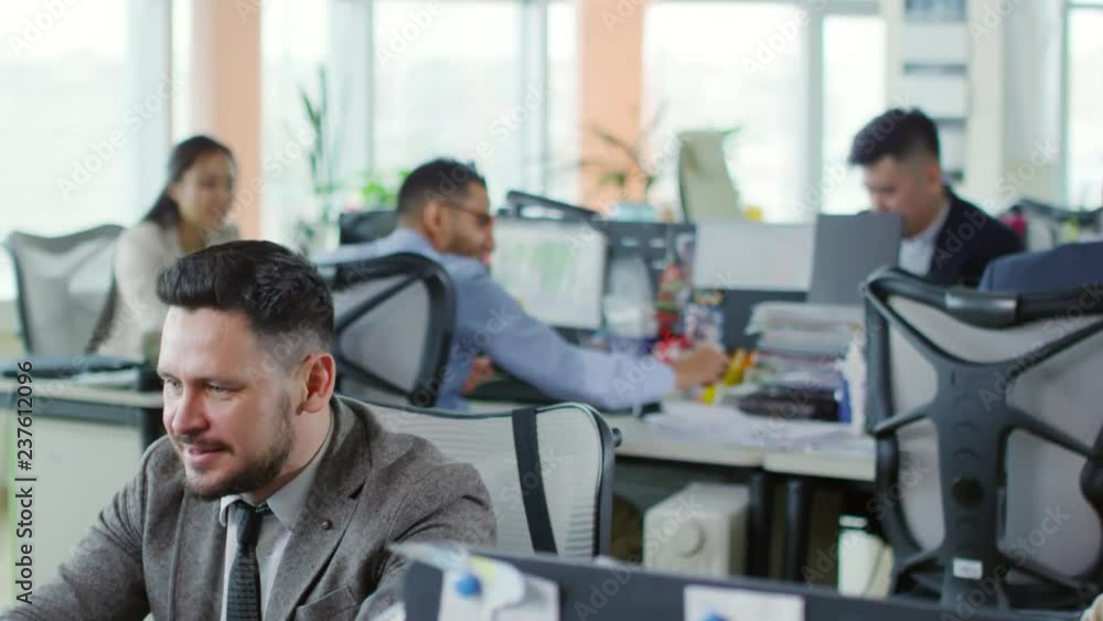 Poster tilt down shot of optimistic middle-aged caucasian man sitting at desk in open-plan office and using