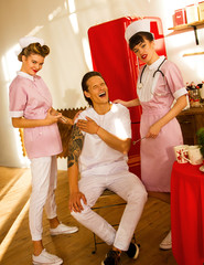 two young stylish nurses give an injection to a man  on the wooden kitchen background with red fridge. emotional male nurse. medical concept. lifestyle