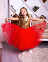 little girl in red shiny dress. kid christmas costume. young girl princess in white room is standing in red fancy skirt  and making kiss with her lips