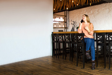 young stylish casual woman with long blonde hair is sitting near wooden table, talking on the mobile phone and waiting somebody with smile in restaurant, free space on white wall, lifestyle concept