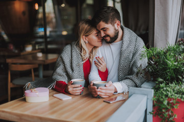 Waist up portrait of charming girl with closed eyes holding hand of her boyfriend while snuggling up to him. They sitting at the table and holding cups of coffee