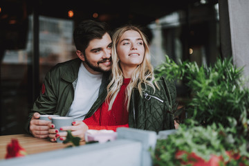 Fototapeta premium Charming girl and her boyfriend sitting at the table and holding cups of coffee. They looking at something interesting and smiling