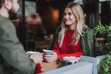 Waist up portrait of charming girl sitting at the table and holding cup of coffee. She looking at bearded man and smiling