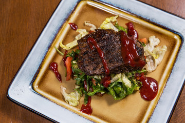 Big meat steak with vegetables, greenery and sauces in square plate on wooden table in luxury restaurant