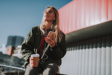 Low angle portrait of charming girl holding cup of coffee while using smartphone and enjoying...