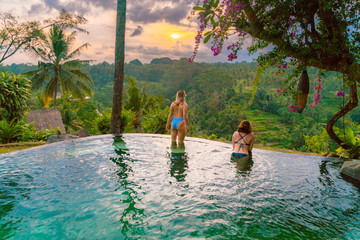 Two women in bathing suits sit on the edge of a blue tropical pool overlooking the jungle