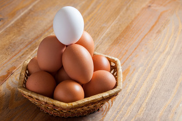 Group of chicken brown eggs and one white lie in a wicker basket on a wooden table.