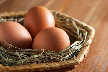 Large brown chicken eggs on straw in a wicker basket on wooden table.