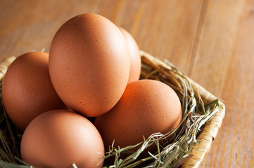 Five brown eggs lay on straw in a wicker basket. Basket on a wooden table. Background out of focus. Close-up.