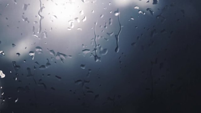Focus on digitally generated raindrops that fall on a foggy window during the day when it rains and the background is blurred.