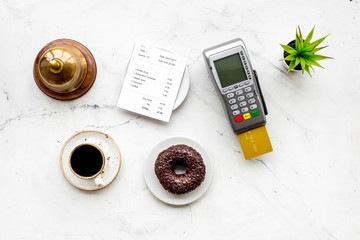 Electronic payments. Pay the bill by card concept. Bank card inserted in payment terminal near bill, service bell, coffee, donut on white stone background top view