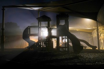 A surreal playground scene on a dark foggy morning with the play equipment backlit by beams of light from the street lamps. Lake Benson Park in Garner North Carolina.