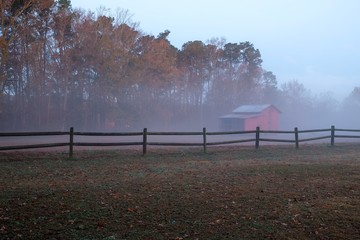 An old red barn behind a wooden fence on a calm foggy morning just before sunrise. Lake Benson Park...