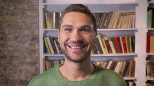 Adult man looking forward, widely smiles. Bookshelves on the background