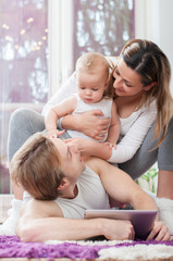 Happy parents laying down on the floor and playing on tablet with their baby boy. Father shows a cartoons to his child.