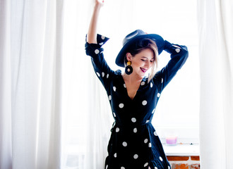 Young redhead girl in black hat and jacket with polka dot dress stay near window.