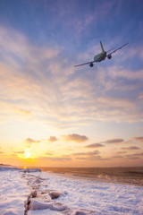 Fototapeta na wymiar Airplane in the sky over the snowy beach and sea at sunset