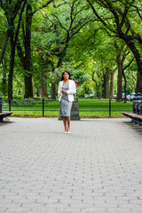 Fototapeta na wymiar Businesswoman Street Fashion. Young Beautiful American Woman traveling in New York, wearing gray patterned off shoulder dress, white coat, light color shoes, walking under green trees at Central Park.