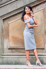Fototapeta premium Young American Woman wearing gray patterned off shoulder dress, light color high heels with ankle strap, wristwatch, standing by old wall at park in New York, raising arm, hand touching cheek, smiling