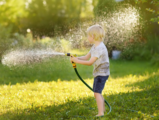 Funny little boy playing with garden hose in sunny backyard. Preschooler child having fun with...
