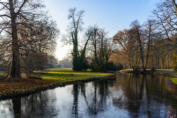 Outdoor sunny tranquil scenery landscape of frozen canal and beautiful waterside in Park Sanssouci surrounded with tree, bush, shrub and winter atmosphere, and background of farther Chinese tea house.