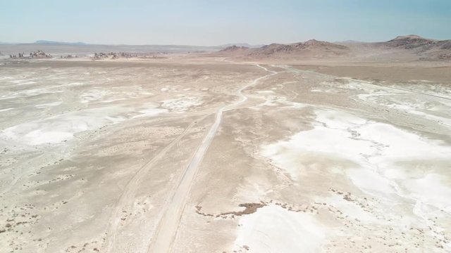 Aerial shot of a dirt road in a desert with white and brown sand. There are brown rocky mountains in the distance 