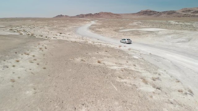 Fast following drone/aerial shot of a car driving in a curvy dirt road in a desert with white sand and rocky mountains in the distance