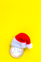 Coin one euro in a red santa claus hat on a yellow background. The concept of financial costs of the European New Year and Christmas. Copy space. Vertical frame