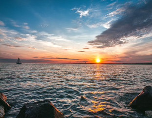 The sailboat sails under the setting sun. Air clouds of different colors. Stone shoreline.