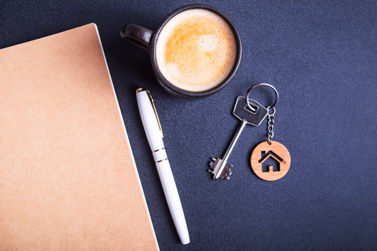 The keys to the new apartment with a wooden key chain house on a black table next to the crafting pad and a cup of coffee. Buying an apartment or house concept. Top view, flat lay