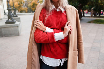 Close up photo of trendy fashionable woman posing in street with blooming spring trees. Wearing Fashion beige jacket and red knitted sweater over white shirt.Female fashion concept. Copy space.