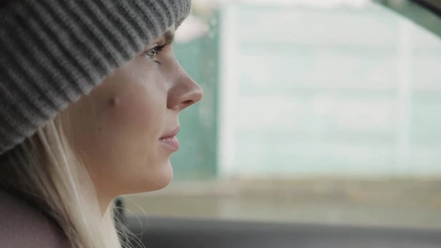 Young adorable blonde woman in grey hat, scarf, blush jacket driving a car, smiling, looking around. Slow motion, cinematic film colors.
