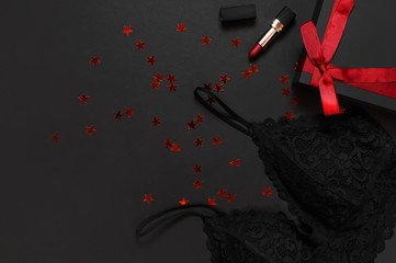 Black gift box with red ribbon, lace bra underwear, red lipstick, holographic confetti on dark background top view flat lay. Female essential erotic accessories, fashionable underwear, gift to woman - obrazy, fototapety, plakaty