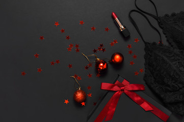 Black gift box with red ribbon, lace bra underwear, red lipstick, Christmas balls, holographic confetti on dark background top view flat lay. Female essential accessories, gift for new year christmas