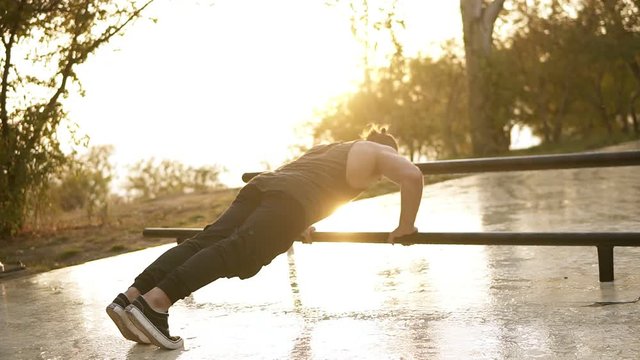 Sport, fitness, young muscular man working out during a workout on the street.Caucasian athlete making push ups on paralel bar. Morning sunshine on the background