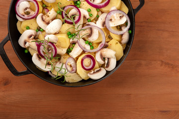 A closeup of a dutch oven with raw vegetables, potatoes, onions, green peas, mushrooms, and rosemary branches, shot from the top on a dark rustic wooden table wih a place for text