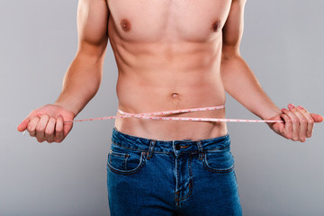 Staying fit. Part of shirtless young man in jeans holding measuring tape while standing against...