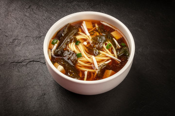 A photo of a bowl of miso soup with tofu, scallions, noodles, wakame, and enoki mushrooms, on a black background with a place for text