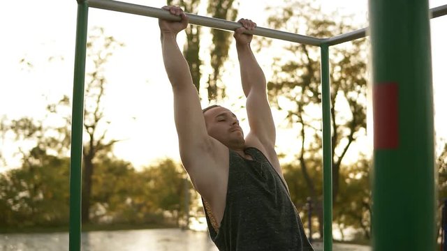 Handsome man having calisthenics training on horizontal bar outdoors. Does gymnastic stunts on the crossbar in the park. Lens flares on the background
