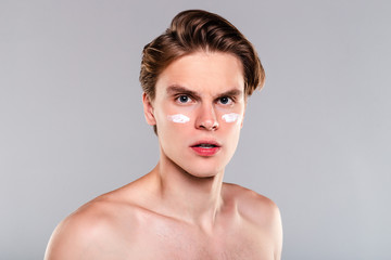 Newer forget about moisturizer. Portrait of handsome shirtless young man with cream on his face and looking at camera while standing against grey background