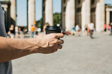 Close-up. A person holds a disposable cup with coffee or another drink while walking or sightseeing in Berlin, Germany. Ahead is the Brandenburg Gate and blurred, unrecognized people.