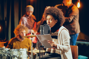 Mixed race woman singing. In background band playing instruments. Home studio interior.