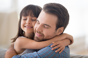 Fototapeta premium Portrait smiling loving handsome Caucasian father and adorable preschool daughter indoors. Daddy and child closed eyes embracing sitting on couch at home. Friendly multi-ethnic family concept