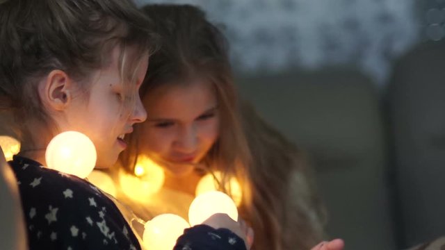 Two girlfriends of the schoolgirl play with luminous garlands in the evening at home. Home illumination, girlish friendship, pajamas party