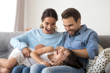 Little funny daughter lying on parents lap while mother father tickling her, laughing have fun play...