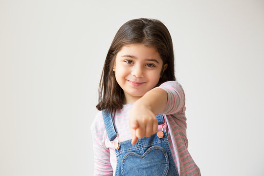 Little Adorable Girl In Denim Overalls Pointing At You. Smiling Cute Child Pointing At Camera With Finger. Isolated On White. Advertisement Concept