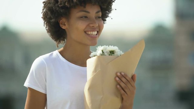 Happy biracial female holding bouquet of white flowers and sincerely smiling