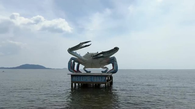Low aerial drone view with camera circling counterclockwise around a giant blue crab statue in coastal Cambodia, Gulf of Thailand. Distant islands and jungle mountains are visible as the camera moves.
