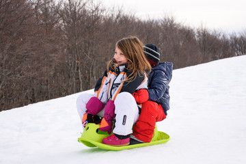 Two children sledding, playing together on snow with slider and laughing